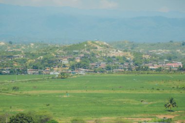 Uzakta küçük bir kasaba olan geniş, açık bir alan. Kasaba yeşil bir manzarayla çevrilidir, bölgeyi ağaçlar ve otlar kaplar kaplar. Gökyüzü açık ve mavidir ve güneş ışıl ışıl parlar.