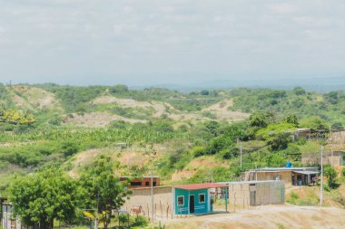 Kırsal alanda küçük yeşil bir ev ve birkaç ev daha var. Gökyüzü bulutlu ve manzara çoğunlukla yeşil.
