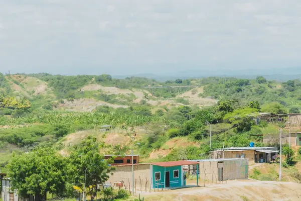 Kırsal alanda küçük yeşil bir ev ve birkaç ev daha var. Gökyüzü bulutlu ve manzara çoğunlukla yeşil.