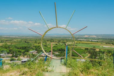 Ortasında sarı bir güneş olan renkli bir yapı. Yapı bir tepenin üzerinde, aşağısında bir kasaba manzaralı.