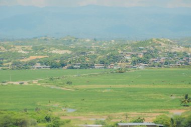 Uzakta küçük bir kasaba olan yemyeşil bir tarla. Gökyüzü açık ve güneş parlıyor.