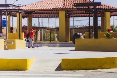 Kırmızı ceketli bir adam sarı bir duvarı temizliyor. Sahne halka açık bir alanda, arka planda bir bank ve küçük bir bina ile çekilmiştir.