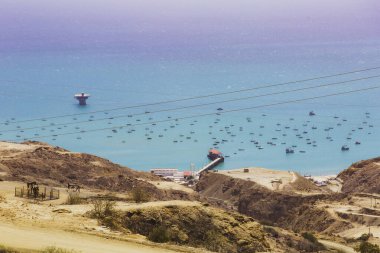 Arka planda büyük bir su kütlesi ve elektrik hattı olan güzel bir plaj sahnesi. Kumsal çeşitli boyutlarda teknelerle dolu, bu da canlı bir atmosfer yaratıyor.