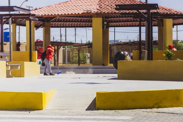 Kırmızı ceketli bir adam sarı bir duvarı temizliyor. Sahne halka açık bir alanda, arka planda bir bank ve küçük bir bina ile çekilmiştir.