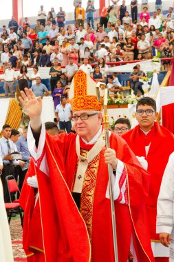 Tumbes, Tumbes, Peru 21-10-2023, Kuvvetlendirme Ayini: Şapkasında beyaz haç olan kırmızı cüppeli bir adam kalabalığa el sallıyor. Adam bir rahip ve kalabalık onu izliyor.
