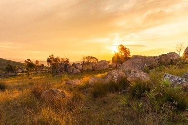 Güneşin arka plandaki manzarası granit kaya parçaları ve kayaların üzerine ve Avustralya 'nın Yeni Güney Galler bölgesindeki Tenterfield yakınlarındaki granit arazideki ağaçların arasına dağılmış bulutlu bir gökyüzünde batıyor..