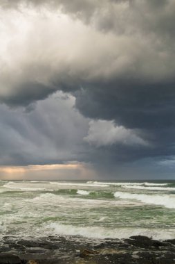 Güçlü fırtına bulutları ve çarpışan dalgalar öğleden sonra güneş ışığıyla birlikte Ballina, Yeni Güney Galler, Avustralya 'daki Shelly Sahili' nde kayalık kıyı şeridi ile buluşur ve gökyüzündeki ve denizdeki ham gücü yakalar..