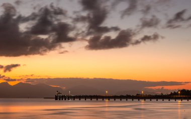 Hinchinbrook Adası üzerinde gün doğumu parıldıyor ve kanalda su yansımaları ve Avustralya 'nın tropik Queensland kentindeki Cardwell şehrinde şafak sökerken ışıkları hala açık olan siluet bir iskele var..