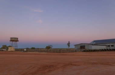 Günbatımının karşı tarafında gökyüzünde mavi bir saat parıldıyor, Avustralya 'nın Quilpie şehrinde yarı kurak bir kırsalda bir çiftlikte yün kırpma kulübesi ve bir su kulesi var..