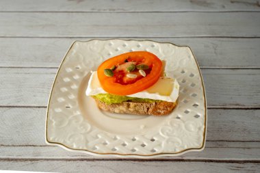 Dekoratif beyaz bir tabakta desenli güzel bir açık yüzlü sandviç sergilenir. Sandviç, üzerinde bir tabaka avokado bulunan bir dilim tam tahıllı ekmekten oluşur.
