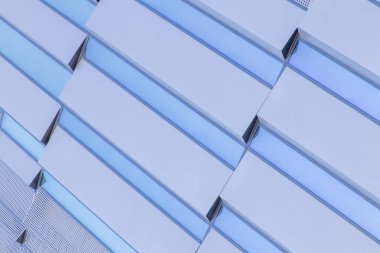 Geometrik metal paneller ve yansıtıcı mavi cam elementlerle modern bir binanın ön cephesinin soyut yakın çekimi. Şablon, çağdaş mimari tasarım, simetri ve minimalizmi vurgular. Yüksek