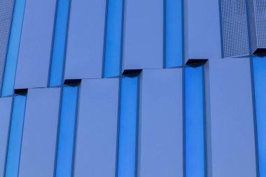 Mavi yansıtıcı cam paneller ve geometrik tasarımla modern bir binanın ön cephesine yakın plan. Soyut mimari modelde çağdaş estetik, simetri ve yenilik sergilenmektedir. Yüksek