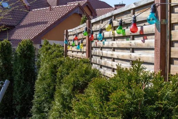 Güneşli bir arka bahçedeki ahşap bir çitte asılı renkli ip ışıkları yeşil çalılar ve kiremitli çatılar. Yüksek kalite fotoğraf