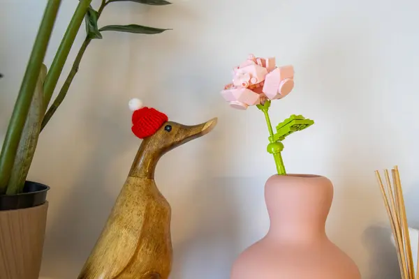 Kırmızı örgü şapkalı tahta ördek heykelciği pastel vazodaki pembe LEGO çiçeğine bakıyor. Estetik minimalist ev dekorasyonu. Doğal ve oyuncu unsurlarla donatılmış. Yüksek kalite fotoğraf