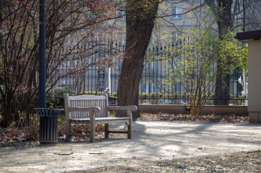Bahar ağaçları, yumuşak güneş ışığı ve gölgeli, kentsel doğa ve siyah demir çitlerle çevrili, sessiz bir park alanında ahşap bir bank..