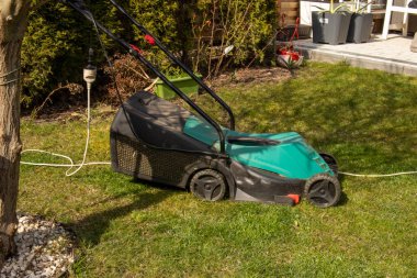 Güneşli bir bahar gününde bahçedeki yeşil çimlerin üzerinde elektrikli çim biçme makinesi. Bahçe bakımı için bahçe malzemeleri hazır..