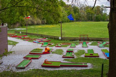 Yeşil park ve ağaçlarla çevrili renkli engellerle dolu açık hava mini golf sahası. Arka planda Avrupa Birliği ve Polonya bayrakları var. Bulutlu bir günde eğlence amaçlı spor alanı.