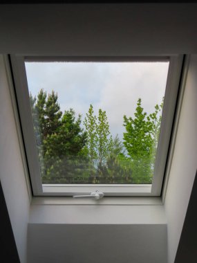 Yeşil ağaçları ve bulutlu gökyüzünü gösteren modern bir çatı penceresinden bak. Minimalist beyaz iç mekan, doğa manzarasını çerçeveliyor..