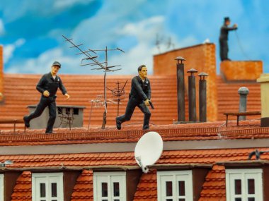 Minyatür polis olay yerini çatıda takip ediyor. Antenler, bacalar ve uydu antenleri arasında hareket halindeler..