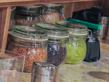 Konforlu bir mutfak veya kafe ortamında ahşap bir rafta çeşitli baharat ve malzemelerle dolu cam kavanozlar..
