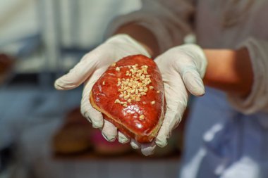 Bir fırın işçisinin yakın plan fotoğrafı, üzerinde doğranmış fındık bulunan jöleli bir pasta tutuyor. Eller eldivende, arka plan hafifçe bulanık.