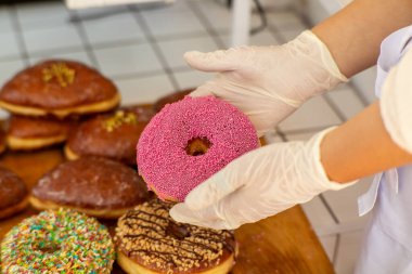 Eldivenli, canlı pembe şekerli bir donut tutuyor. Fırında ya da dükkan ortamında renkli pastalar. Sokak yemeği ya da tatlı konsepti.
