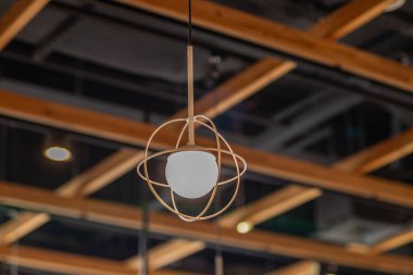 Modern asılı lamba, geometrik metal çerçeve ve beyaz ampul, ahşap ızgara tavana dayanıyor. Modern iç aydınlatma stili.
