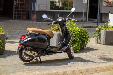 Eski model siyah scooter, bej koltuklu, parke taşlı bir caddede park halinde, bir kafe ve yeşil bitkilerin yanında. Güneşli bir günde klasik şehir ulaşımı.
