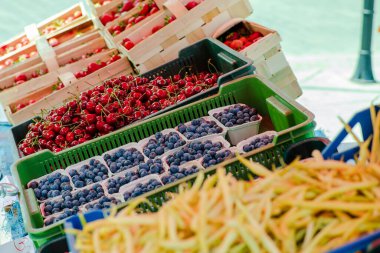 Tepsiler dolusu yaban mersini, kiraz ve sarı fasulyeli renkli market tezgahı. Canlı gün ışığında yakalanan taze mevsimlik meyve ve sebzeler. Gıda ve tarım temaları için mükemmel.