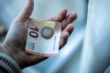 Bir kişi elinde 10 avroluk banknot ve 2 avroluk para tutuyor. Arka plan hafifçe bulanık, dikkatini para birimine odaklıyor..
