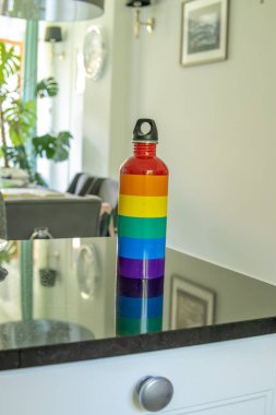 Gökkuşağı desenli paslanmaz çelik bir su şişesi parlak, rahat bir odada yansıtıcı bir yüzeyde duruyor. Modern bir iç mekanda LGBTQ destek sembolü.