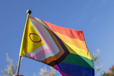 Açık mavi gökyüzüne karşı gün ışığında açık havada uçan cinsiyetler arası sembolün yer aldığı İlerleme Gururu Bayrağı. Katılım, çeşitlilik ve LGBTQ haklarının canlı bir sembolü.