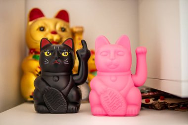 İki Maneki-neko figürü: biri siyah, orta parmağı kalkık ve diğeri de klasik pozlu neon pembeli. Şanslı kedi geleneğini eğlenceli, alaycı bir şekilde ele alalım..