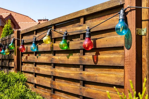 Bahçedeki ahşap bir çitte asılı renkli ip ışıkları. Şenlikli açık hava dekorasyonu, günışığında klasik tarzda ampullerle, neşeli bir atmosfer yaratıyor..