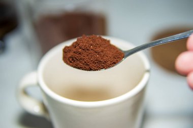 Beyaz bir seramik kupanın üzerinde duran bir kaşık dolusu kahve. Temiz bir mutfak arka planında sabah kahvesi hazırlama görüntüsü..