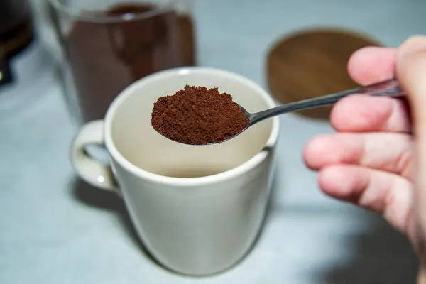 Bir kaşık dolusu kahveyi seramik bardağın üstünde tutan bir el. Parlak bir mutfak ortamında kahve yapma sürecinin yakın görüntüsü.