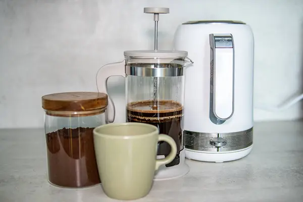 Kahve hazırlama seti, bir kavanoz öğütülmüş kahve, bir Fransız presi, modern bir elektrikli su ısıtıcısı, ve açık mutfak yüzeyinde yeşil bir kupa..