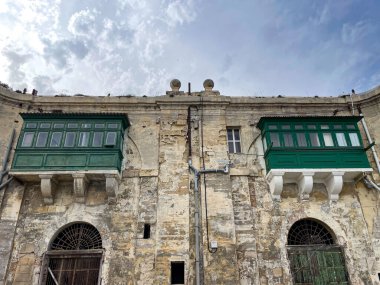 Malta 'daki eski taş bina yıpranmış duvarlar ve iki geleneksel kapalı ahşap balkon parçalı bulutlu bir gökyüzünün altında yeşile boyanmış..