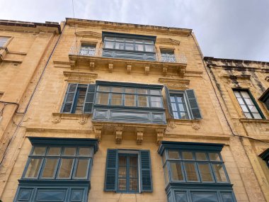Geleneksel Malta cephesi mavi ahşap balkonları ve mavi gökyüzünün altındaki sarı taş bir binanın kepenkleri. Dikey mimari görünümü.