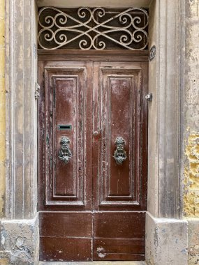 Süslü metal ızgaralı klasik ahşap kapı ve yıpranmış taş bir cephede aslan başlı tokmaklar. Malta 'da tarihi giriş.