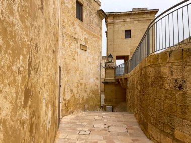 Mdina, Malta 'da, yıpranmış taş duvarlar ve üzerinde metal bir korkuluk ile çerçevelenmiş, sakin, altın renkli bir sokak. Sıcak tonlar ve dokular yüzyıllardır süren Akdeniz tarihini yansıtıyor..