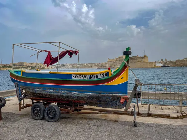 Limandaki bir karavanda dinlenen San Pawl adlı geleneksel Malta luzzu teknesi, Vallettas silueti ve arka planda sakin deniz parçalı bulutlu bir gökyüzünün altında.