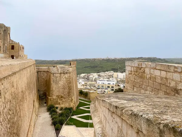 Dar bir yolu olan taş kale duvarları ve aşağıdaki manikürlü yeşil bahçe Malta 'da sisli bir gökyüzünün altındaki uzak bir kasabaya bakıyor..