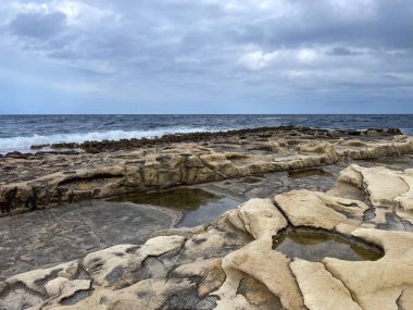 Bulutlu bir gökyüzünün altında kireç taşı oluşumları ve gelgit havuzlarıyla adaların doğal ham güzelliğini yakalayan kayalık bir Malta kıyısı..