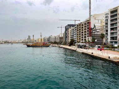 Sliema, Malta 'daki modern binaların yakınında durgun turkuaz sularda geleneksel yelkenli gemi. Güneşli bir günde şehir limanı ve deniz kontrastı.
