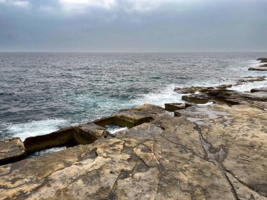 Malta 'daki kayalık sahil şeridi dramatik taş oluşumları ve kasvetli bir gökyüzü altında dalgaları çarpıştırıyor. Doğal deniz manzarası Akdeniz kıyılarının saf güzelliğini gösteriyor..