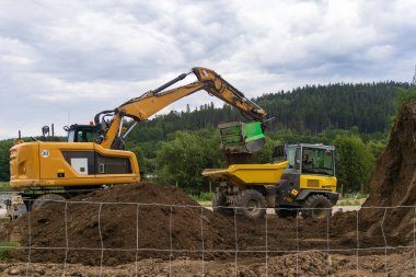 Kazıcı, arka planında ormanlık tepeler olan bir inşaat alanında sarı bir çöp kamyonuna toprak yüklüyor. Bulutlu gökyüzü altında yeryüzü çalışmaları devam ediyor.