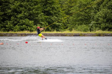 Wakeboardcu göl kıyısına yakın bir yere gider ve arka plan olarak yoğun yeşil bir orman ile küçük bir sıçrama yaratır. Atış hareketi ve dengeyi yakalar..