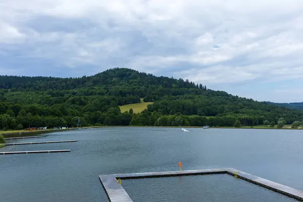 Rıhtımı olan huzurlu bir göl ve bulutlu bir gökyüzünün altında yemyeşil bir tepe. Sakin suları ve orman yamaçlarıyla sakin bir doğal manzara..