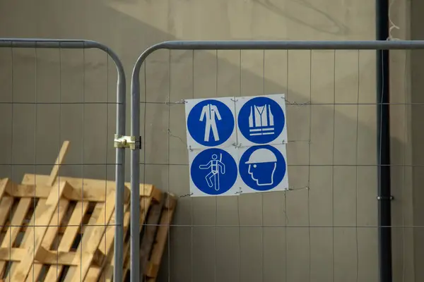 İnşaat çitlerindeki mavi güvenlik işaretleri koruyucu ekipman gerektirdiğini gösteriyor: tulum, yelek, kask, kulak koruyucu ve eldiven. Tahta paletler yakınlarda..